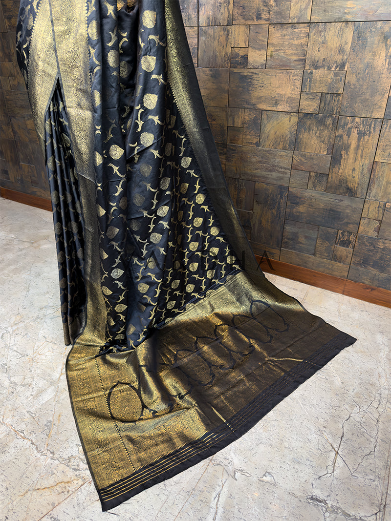 Black and gold patterned fabric draped over a wooden surface. Elegant Black festive Banarasi Satin Silk Saree from House of Vardha with radiant Gold Zari work, perfect for evening wear and special occasions. We offer Ready to wear, Pre drape service, Saree Add on, Custom Stitched Blouse, Fall and Edging, Gift Packaging at Complimentary Price!