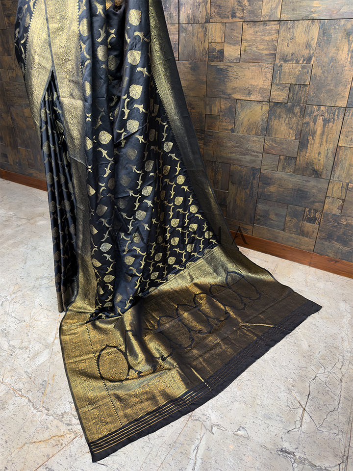 Black and gold patterned fabric draped over a wooden surface. Elegant Black festive Banarasi Satin Silk Saree from House of Vardha with radiant Gold Zari work, perfect for evening wear and special occasions. We offer Ready to wear, Pre drape service, Saree Add on, Custom Stitched Blouse, Fall and Edging, Gift Packaging at Complimentary Price!