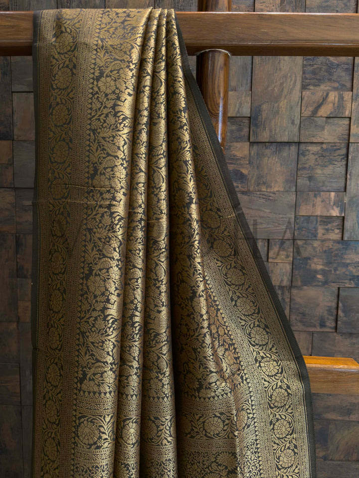 Textured fabric draped over a wooden chair against a wooden paneled wall.  Close-up of zari work on black Mysore silk saree featuring floral vines and creeper patterns
