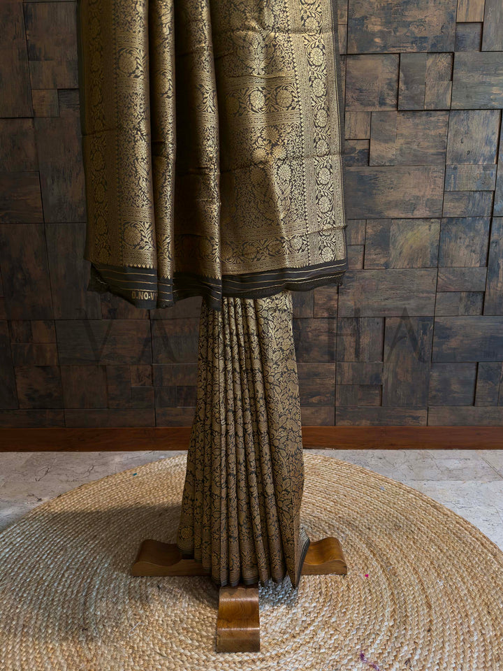 Decorative curtain with intricate patterns on a wooden stand against a textured wall.  Black Mysore silk saree pleats highlighted with intricate zari motifs and golden weaving
