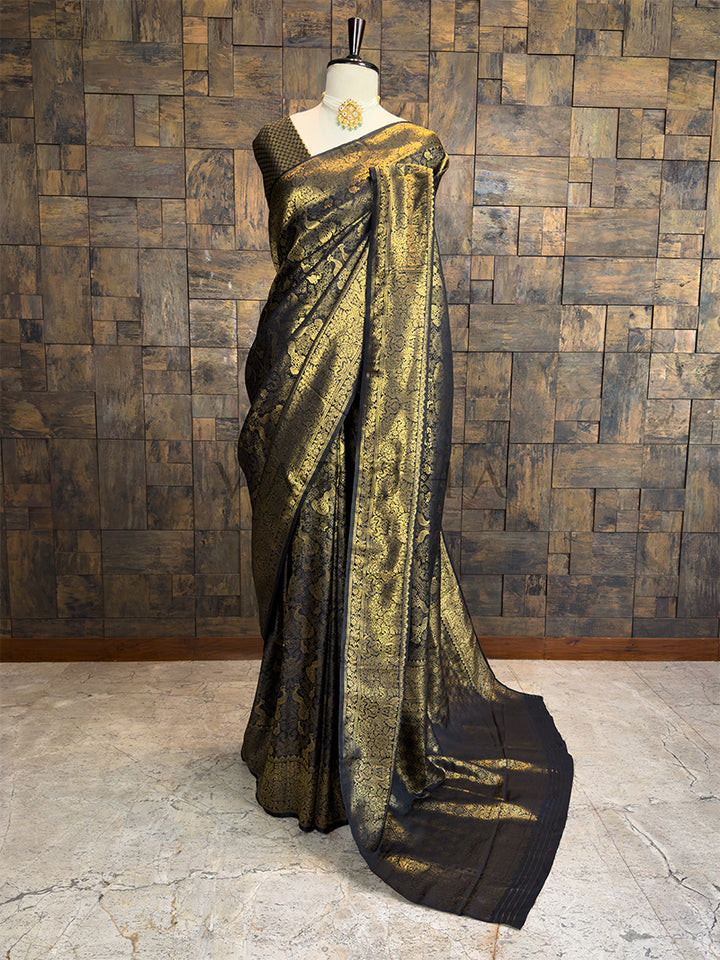 Black and gold saree on a mannequin against a wooden paneled wall. House of Vardha premium black silk saree with heavy gold zari peacock motifs and rich brocade border, draped on a mannequin.