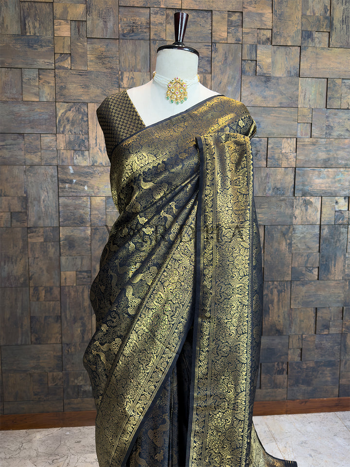 Black and gold saree on a mannequin against a wooden paneled wall. Elegant dark and gold woven saree by House of Vardha featuring intricate traditional peacock and floral zari craftsmanship.