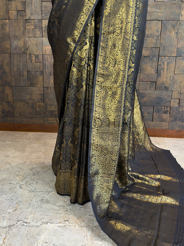 Decorative black and gold fabric draped over a wooden panel wall. Close-up of House of Vardha black festive saree showcasing elaborate gold zari detailing along the pallu and wide traditional border.