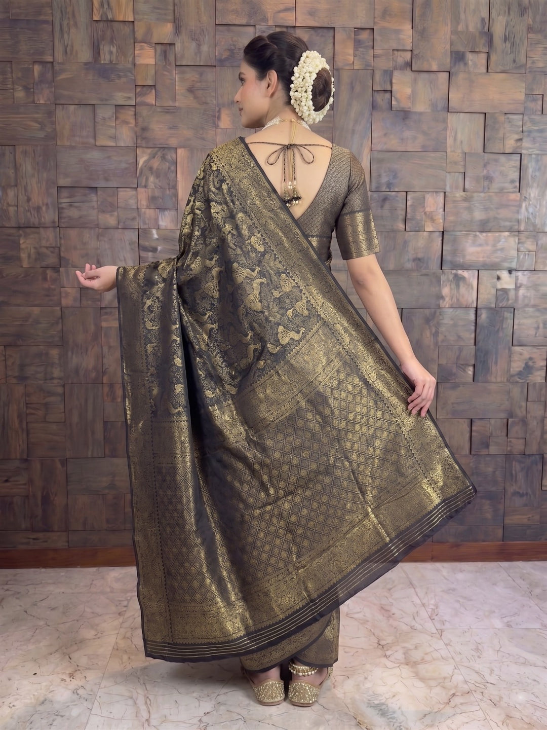 A woman wearing a metallic bronze and gold brocade saree with a matching blouse, standing in front of a rustic wooden paneled wall. A full-length back view of a woman draped in a black Kanjivaram silk saree, holding the edge of the pallu out with her right hand to display the elaborate gold zari weaving. She is wearing a matching black patterned blouse with a deep V-neck back design featuring string ties and decorative tassels.