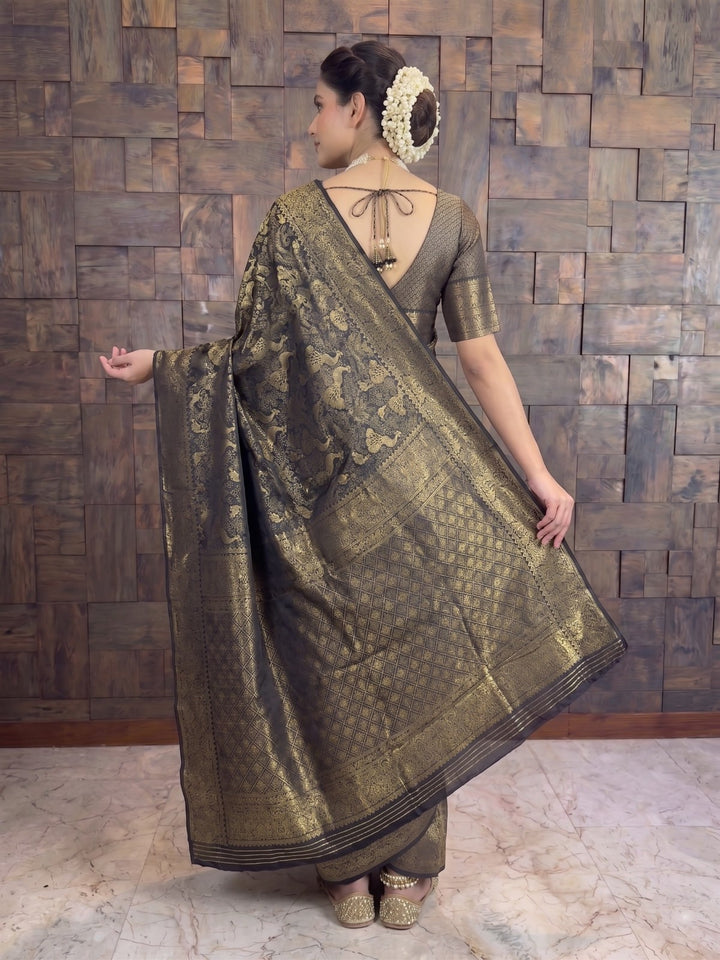 A woman wearing a metallic bronze and gold brocade saree with a matching blouse, standing in front of a rustic wooden paneled wall. A full-length back view of a woman draped in a black Kanjivaram silk saree, holding the edge of the pallu out with her right hand to display the elaborate gold zari weaving. She is wearing a matching black patterned blouse with a deep V-neck back design featuring string ties and decorative tassels.