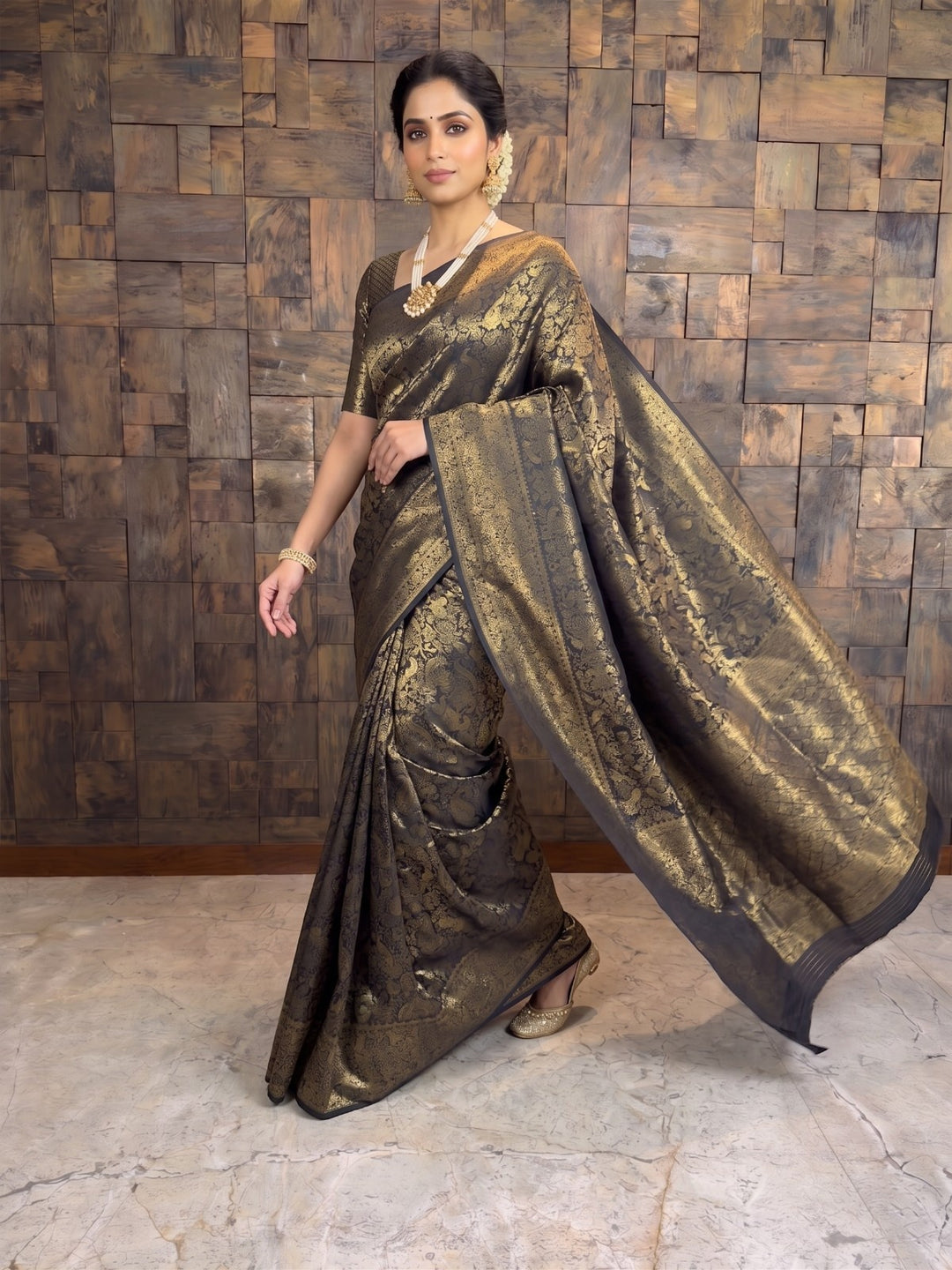 A woman modeling a dark brown and gold brocade saree with a wide draped pallu, standing against a rustic wooden paneled wall. A full-length dynamic shot of a woman stepping forward in a black Kanjivaram silk saree. The graceful movement showcases the rich gold zari details across the fabric and the elegant sway of the pallu cascading behind her as she walks.