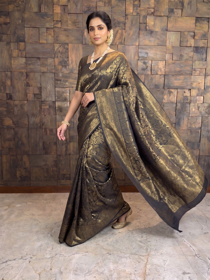 A woman modeling a dark brown and gold brocade saree with a wide draped pallu, standing against a rustic wooden paneled wall. A full-length dynamic shot of a woman stepping forward in a black Kanjivaram silk saree. The graceful movement showcases the rich gold zari details across the fabric and the elegant sway of the pallu cascading behind her as she walks.