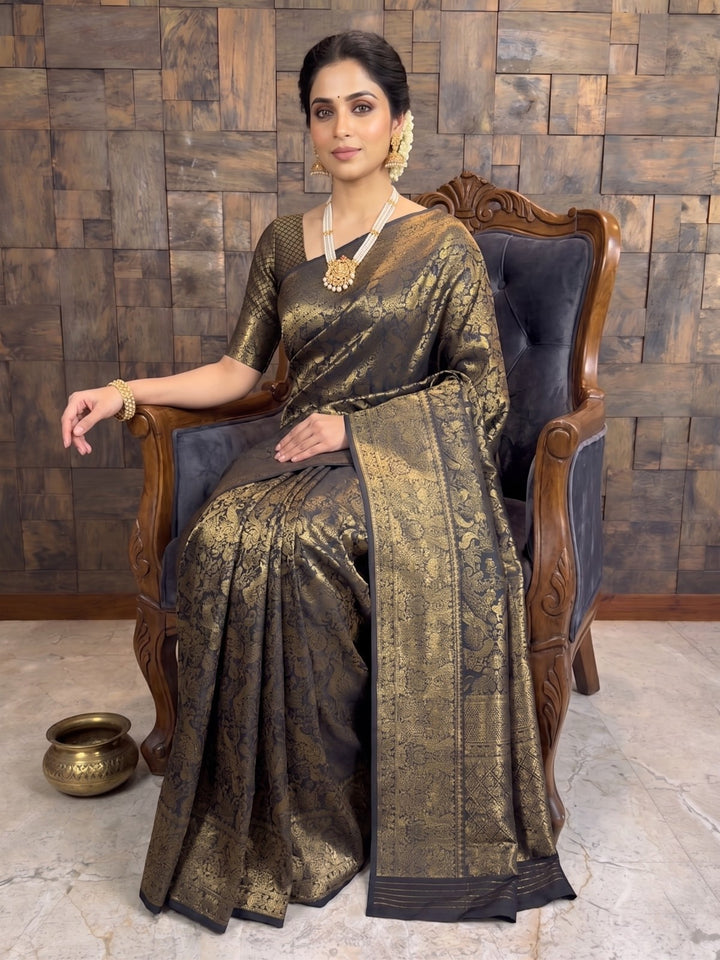 A woman seated in an ornate wooden armchair wearing a dark metallic bronze saree with gold zari embroidery and a wide border, styled with a matching blouse, necklace, and earrings against a rustic wood-paneled wall. A full-length shot of a woman seated gracefully on a dark, carved wooden armchair with plush upholstery. She is dressed in the black Kanjivaram silk saree with heavy gold zari, resting her right arm on the armrest, while a small decorative brass pot sits on the floor near her feet.