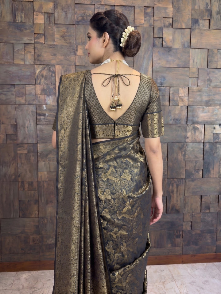 A woman shown from the back wearing a dark metallic bronze and gold patterned saree with a matching embroidered blouse, standing against a rustic wooden wall. A detailed back view focusing on the upper half of a woman wearing a black Kanjivaram silk saree. The close-up highlights her braided bun adorned with a white gajra, and the matching blouse&