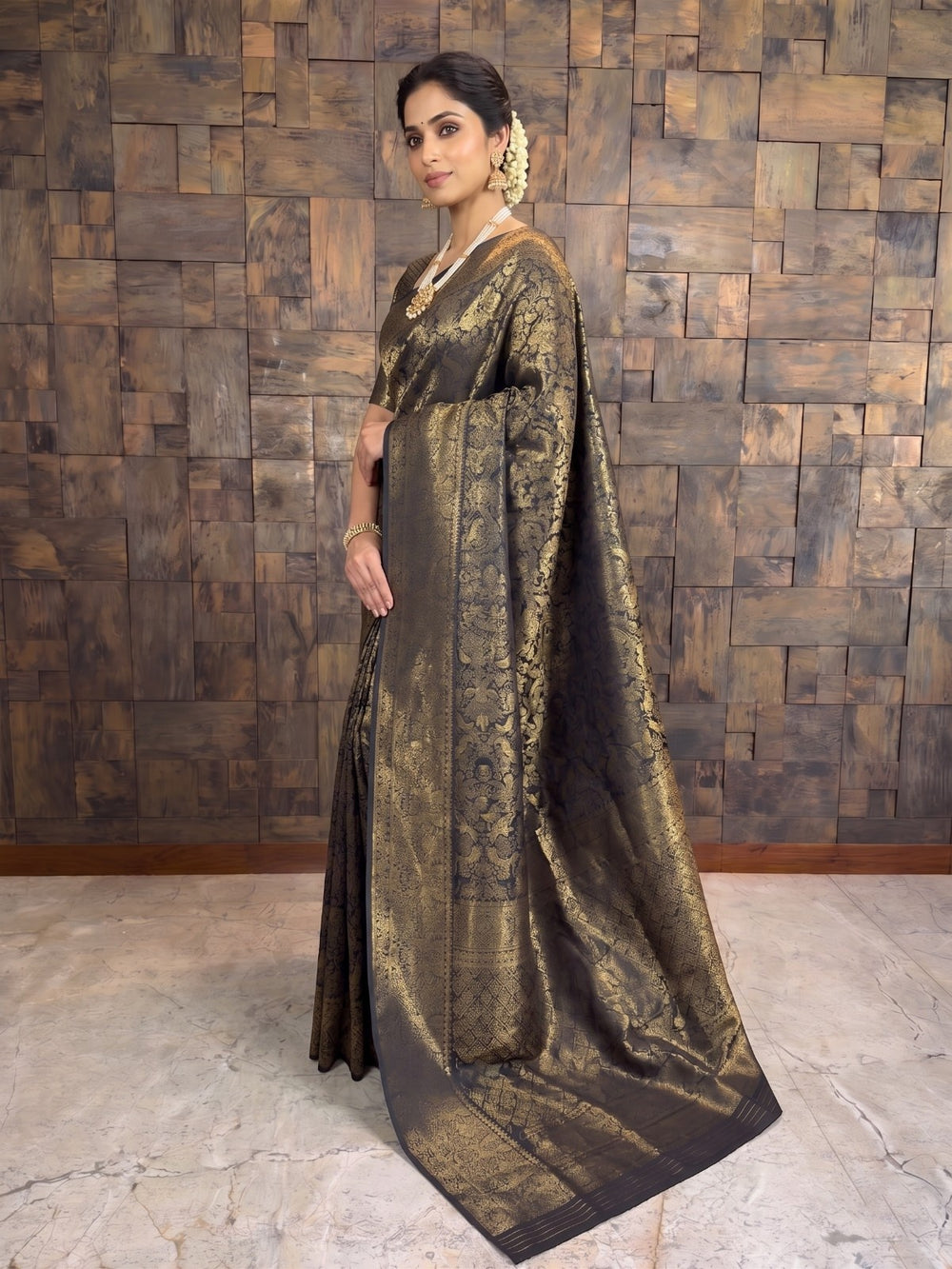 A woman models a dark brown and gold brocade saree in front of a rustic wooden paneled wall. The saree features intricate gold zari embroidery and a wide border, with a matching dark blouse. A full-length side profile view of a woman wearing a black and gold zari Kanjivaram silk saree. The angle highlights the intricate gold motifs woven throughout the dark fabric and the thick zari border beautifully draped over her left shoulder.