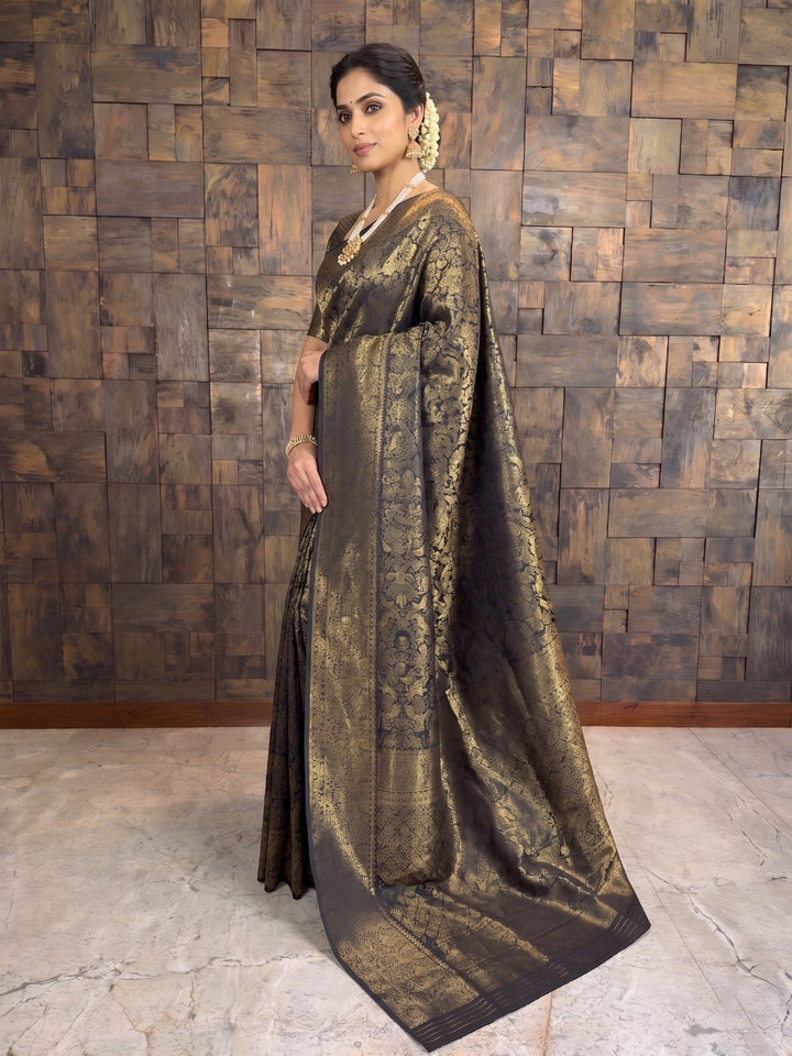 A woman models a dark brown and gold brocade saree in front of a rustic wooden paneled wall. The saree features intricate gold zari embroidery and a wide border, with a matching dark blouse. A full-length side profile view of a woman wearing a black and gold zari Kanjivaram silk saree. The angle highlights the intricate gold motifs woven throughout the dark fabric and the thick zari border beautifully draped over her left shoulder.