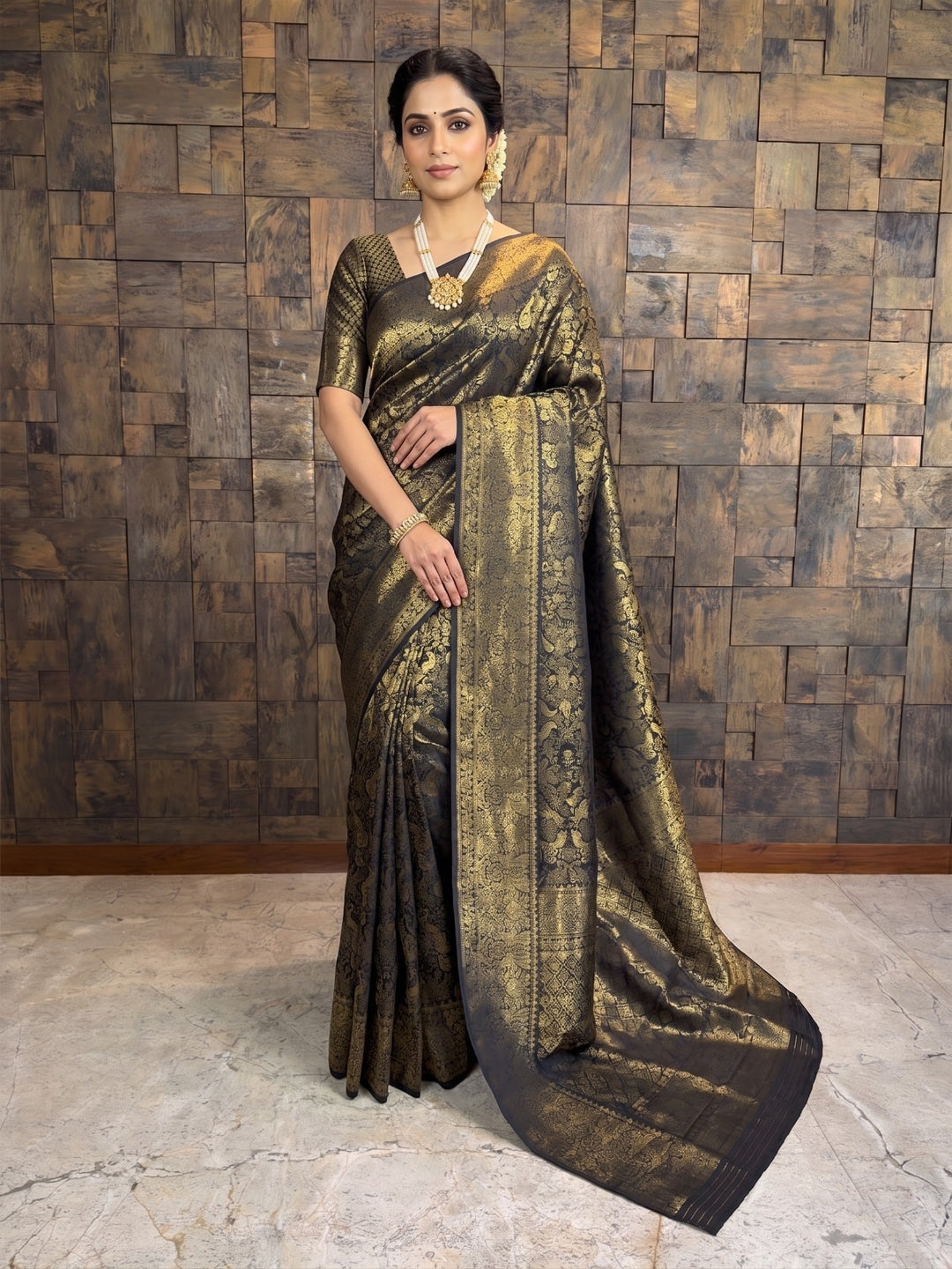 A woman models a black and gold zari woven saree with a matching blouse, standing against a rustic wooden paneled wall. A full-length front view of a woman standing in a black Kanjivaram silk saree adorned with heavy gold zari woven floral and traditional patterns. She wears a matching short-sleeved blouse, a prominent pearl and gold necklace, gold earrings, and a white floral gajra in her elegantly styled hair.