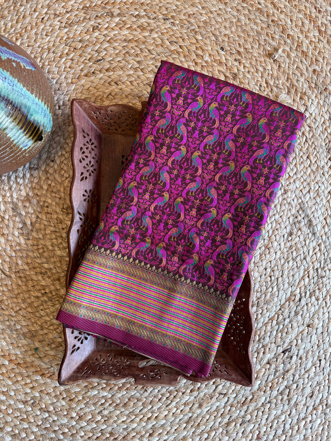 Folded black satin Tanchoi silk saree with colorful thread-woven parrot motifs and golden striped border, elegant for gifting or pooja