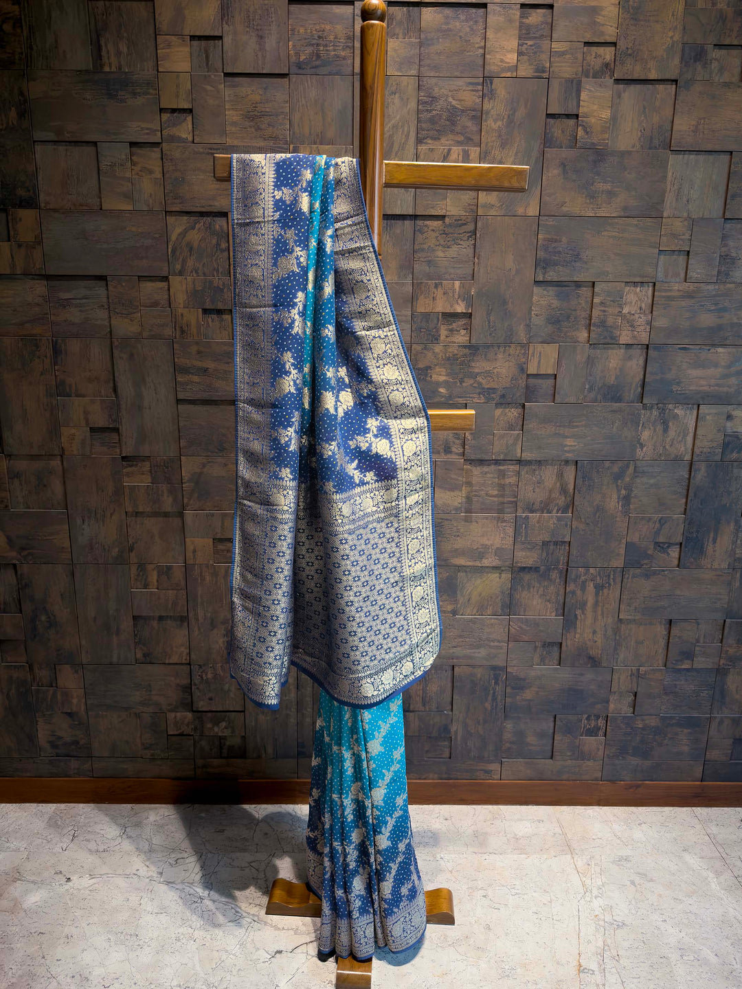 Blue and white patterned saree on a wooden stand against a textured brown wall.