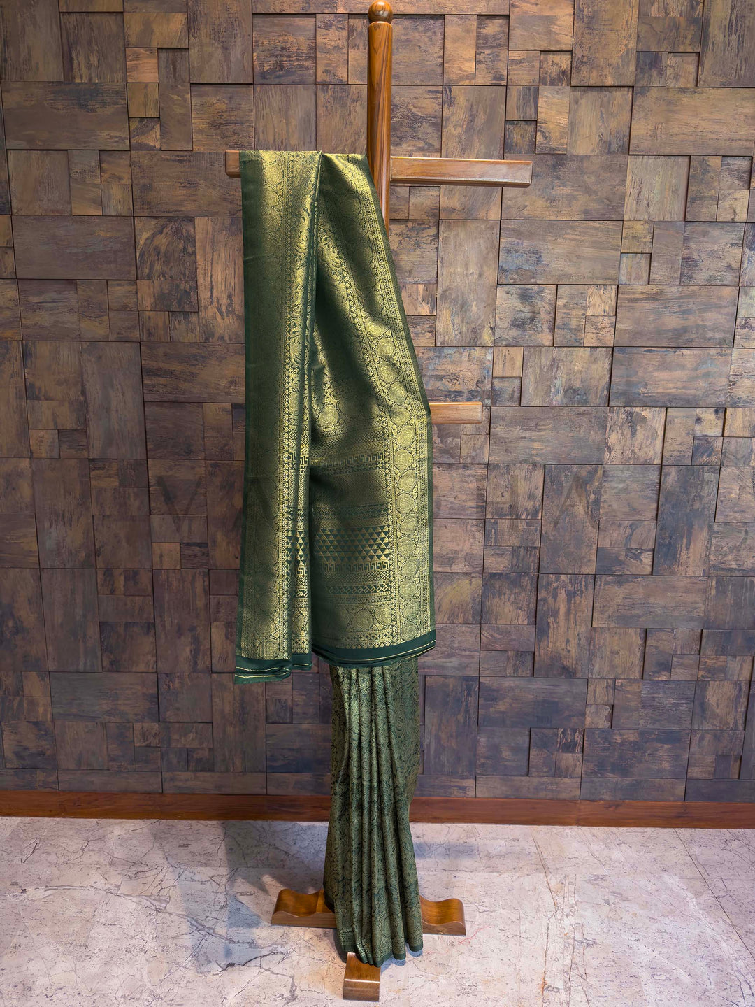 Green textured fabric draped over a wooden hanger against a wooden paneled wall. Bottle Green Kanjivaram Silk Saree with antique gold zari floral motifs displayed on hanger