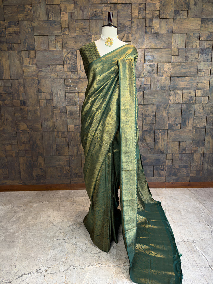 Green saree on a mannequin against a wooden paneled wall. House of Vardha Bottle Green Golden Zari Kanjivaram Silk Saree featuring heavy traditional gold woven motifs, draped on a mannequin.