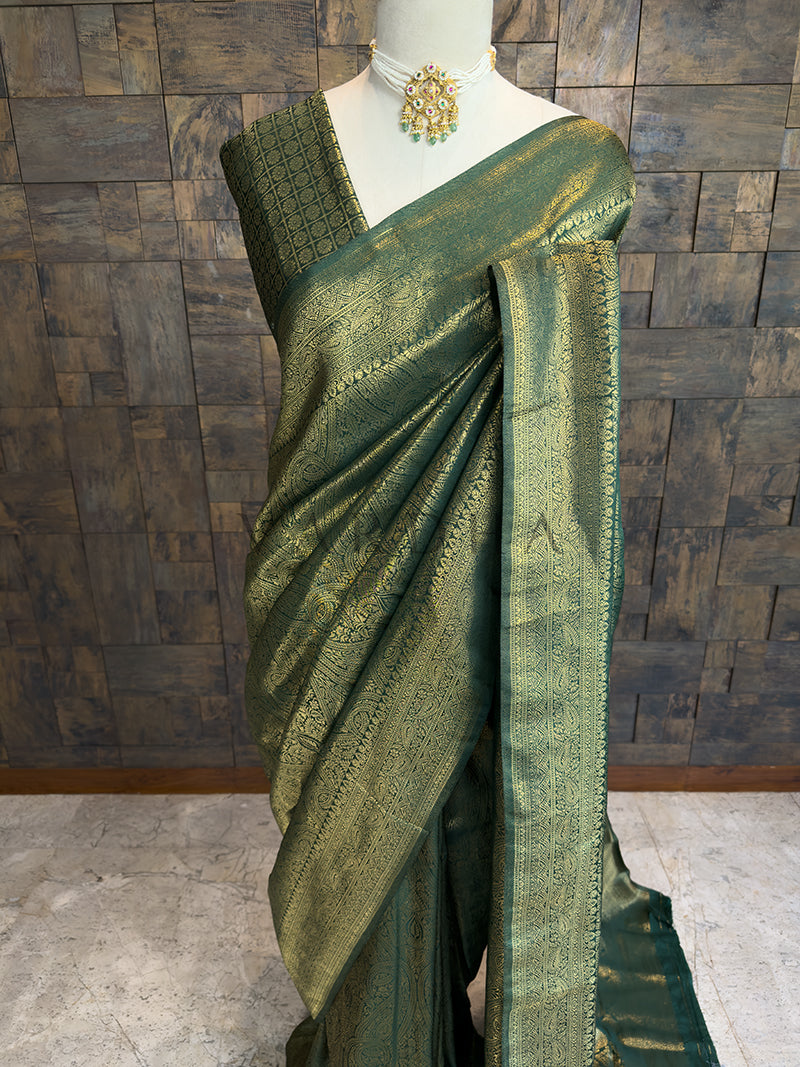 Green saree with gold border on a mannequin against a wooden paneled wall. Premium dark green Kanjivaram bridal saree by House of Vardha showcasing dense gold zari floral and paisley weaving.