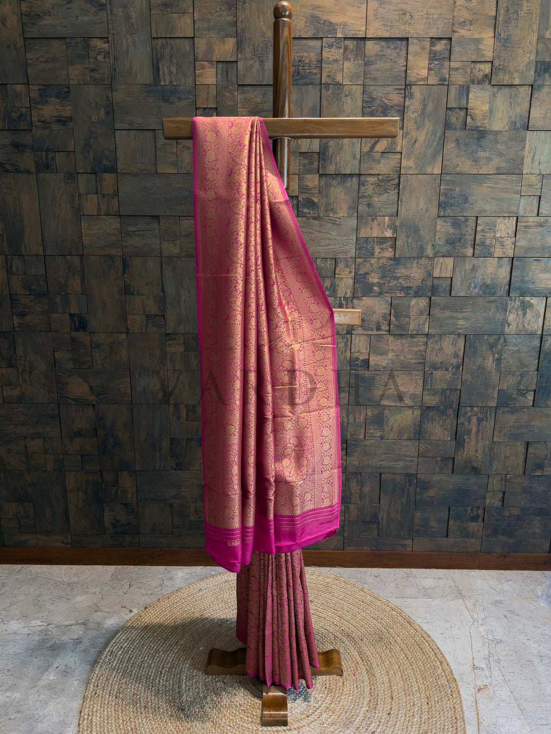 Pink saree with a border draped over a wooden stand against a textured wall. Pink Mysore Silk Saree with golden zari floral weaving on display