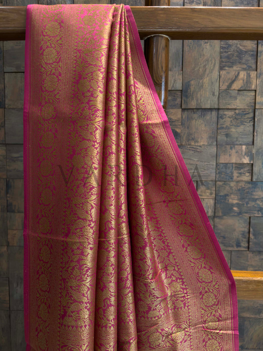Pink and gold saree draped over a wooden rod against a stone wall background Close-up of zari floral motifs on bright pink viscose silk saree