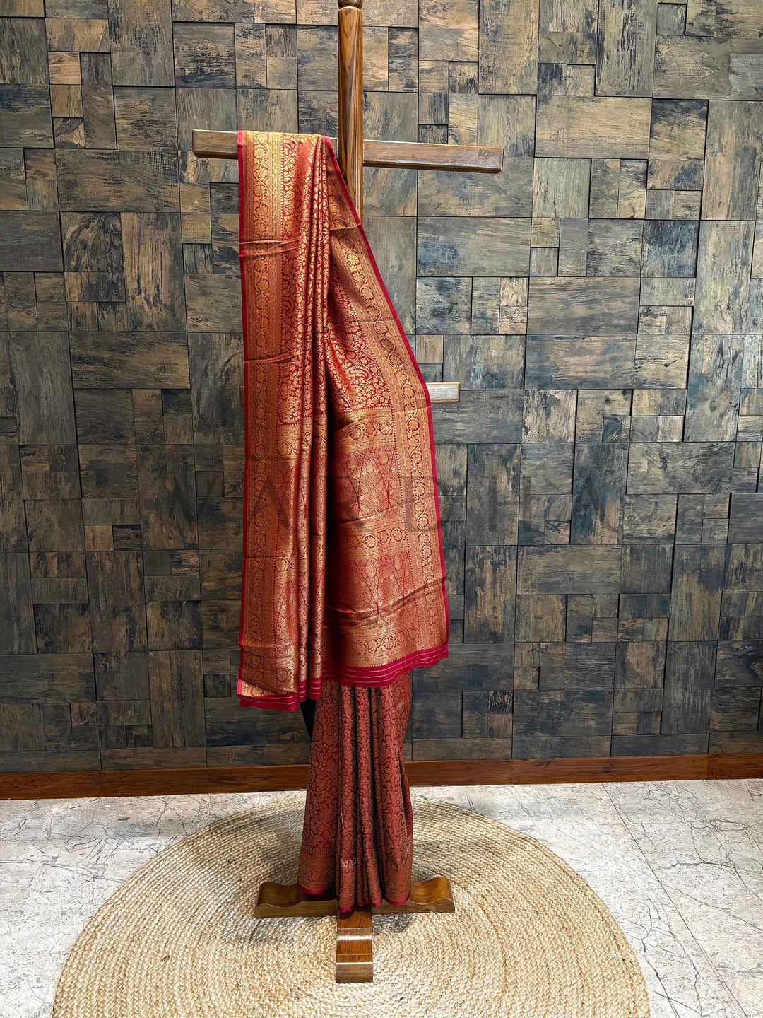 Red saree with gold patterns on a wooden stand against a textured wall. Wine Maroon Mysore silk saree in pure viscose fabric with golden zari floral vines and broad border showcased on mannequin.