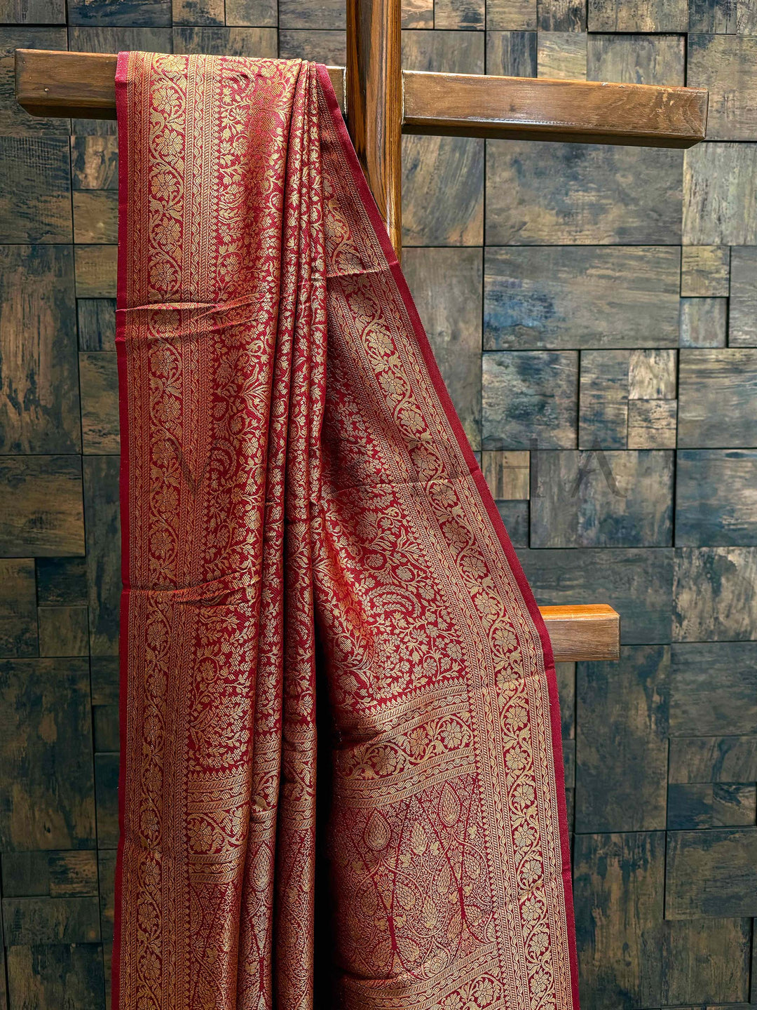 Red patterned fabric draped over a wooden cross against a stone wall. Close-up of intricate golden zari weaving with paisley and leaf motifs on the wine maroon Mysore silk saree