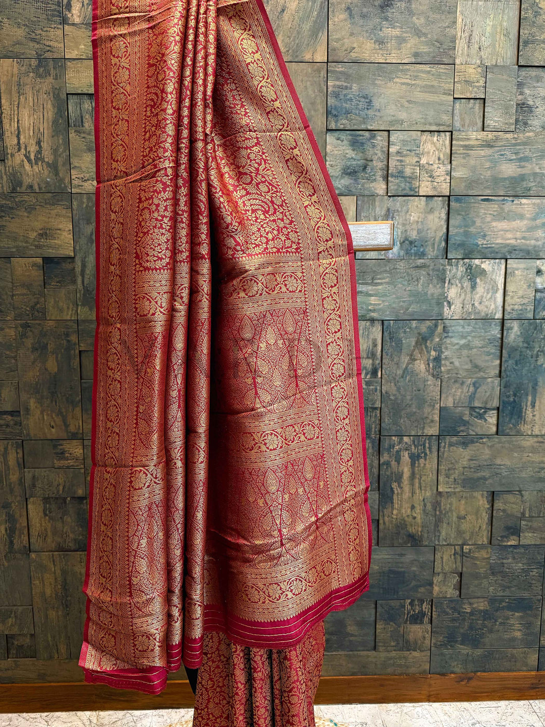 Red saree with intricate patterns draped over a wooden surface against a textured wall. Detailed view of the pallu featuring temple-style jaal design in metallic gold over rich viscose base