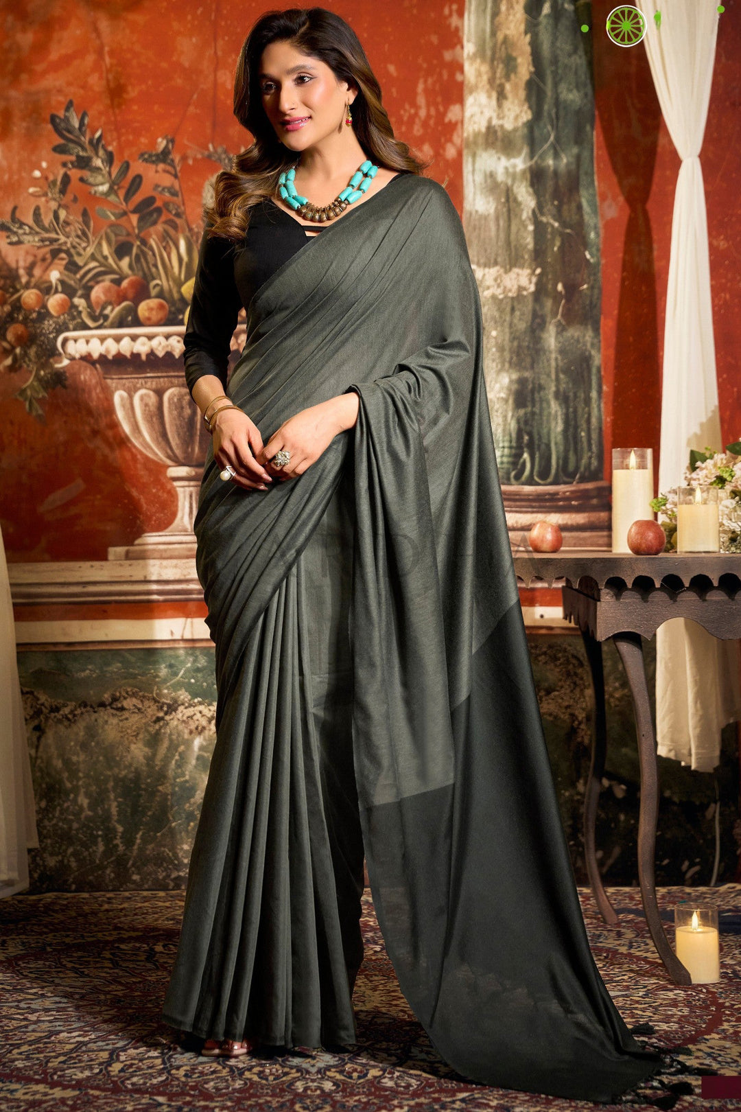 Woman in a dark green saree standing in an ornate room with decorative elements. Full front view of a woman wearing a sophisticated charcoal grey mal cotton saree featuring a bold black contrast pallu and fancy tassels.
