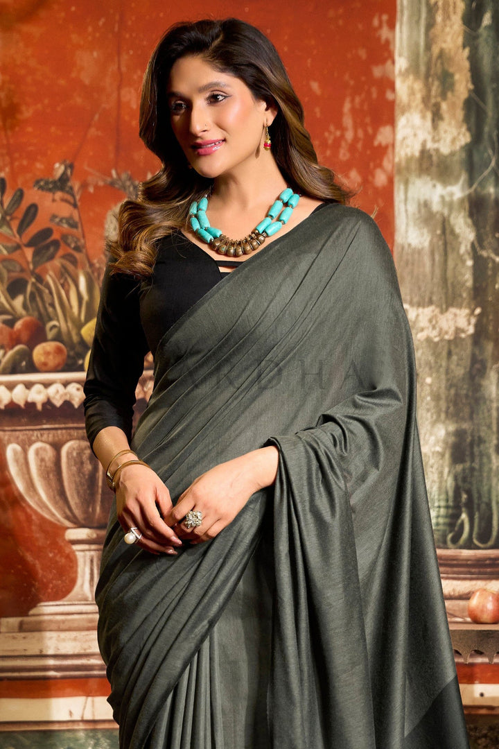 Woman wearing a dark green saree with a textured wall background Detailed fabric close-up of the charcoal grey mal cotton saree highlighting the perfectly pleated texture and the stark black contrast pallu edge with tassels. Close-up of a beautiful woman draped in a soft charcoal grey mal cotton saree, showcasing the black contrast blouse and elegant summer office wear look.