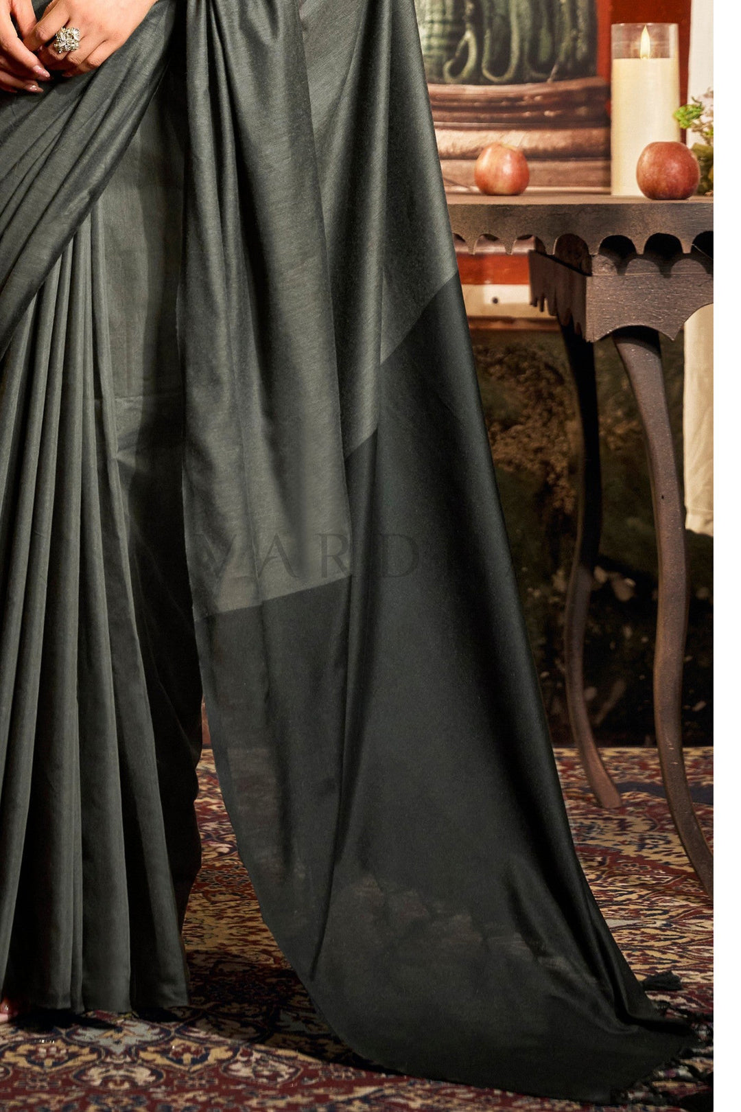 Dark green fabric draped over a chair with a blurred background of a room. Detailed fabric close-up of the charcoal grey mal cotton saree highlighting the perfectly pleated texture and the stark black contrast pallu edge with tassels.