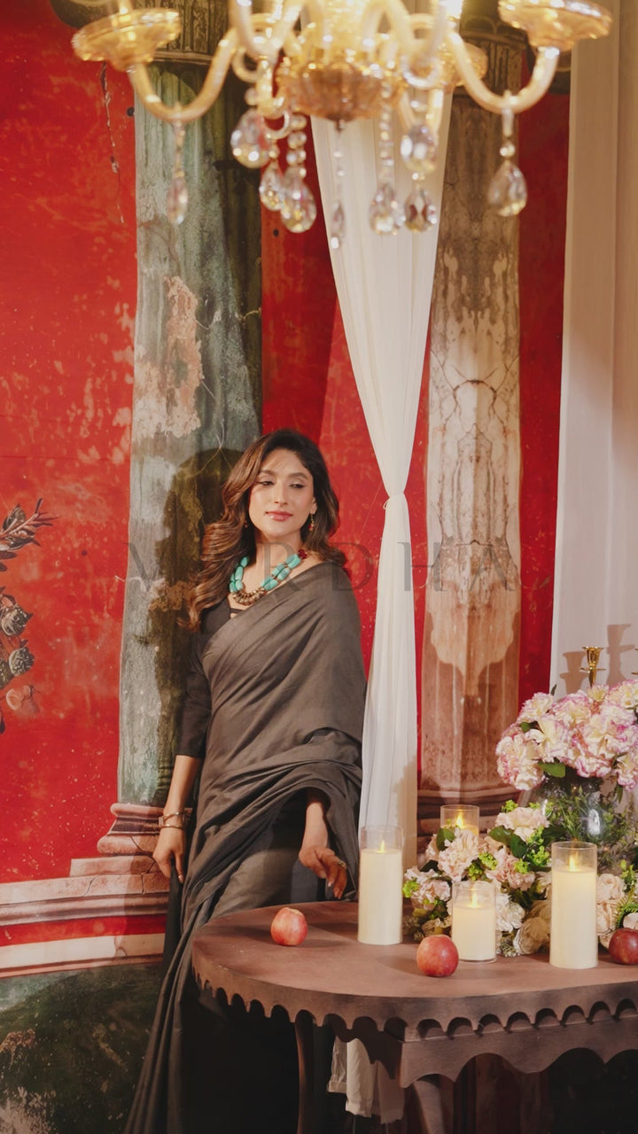 A woman wearing a dark brown draped saree stands indoors beside a decorated table with lit candles, flowers, and red apples, set against a red wall and ornate drapery. Beautiful woman posing gracefully in an elegant grey and black dual-tone mal cotton saree, highlighting its minimalist and effortlessly chic aesthetic.