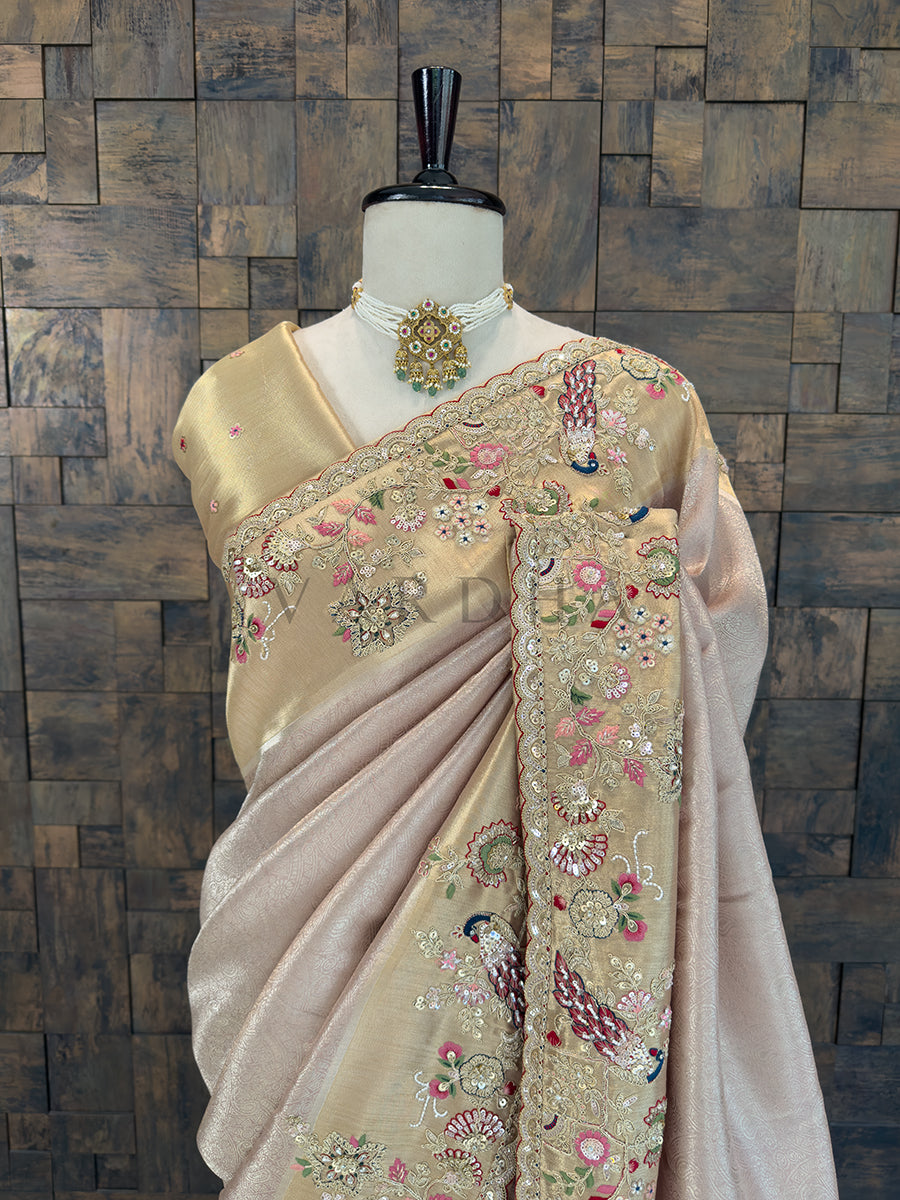 Beige saree with floral embroidery on a mannequin against a wooden paneled wall. Close-up of a luxurious rose gold party-wear saree showing detailed zari, sequin, and thread work in floral and peacock motifs on the shoulder pallu, accessorized with a traditional pearl and kundan choker necklace.
