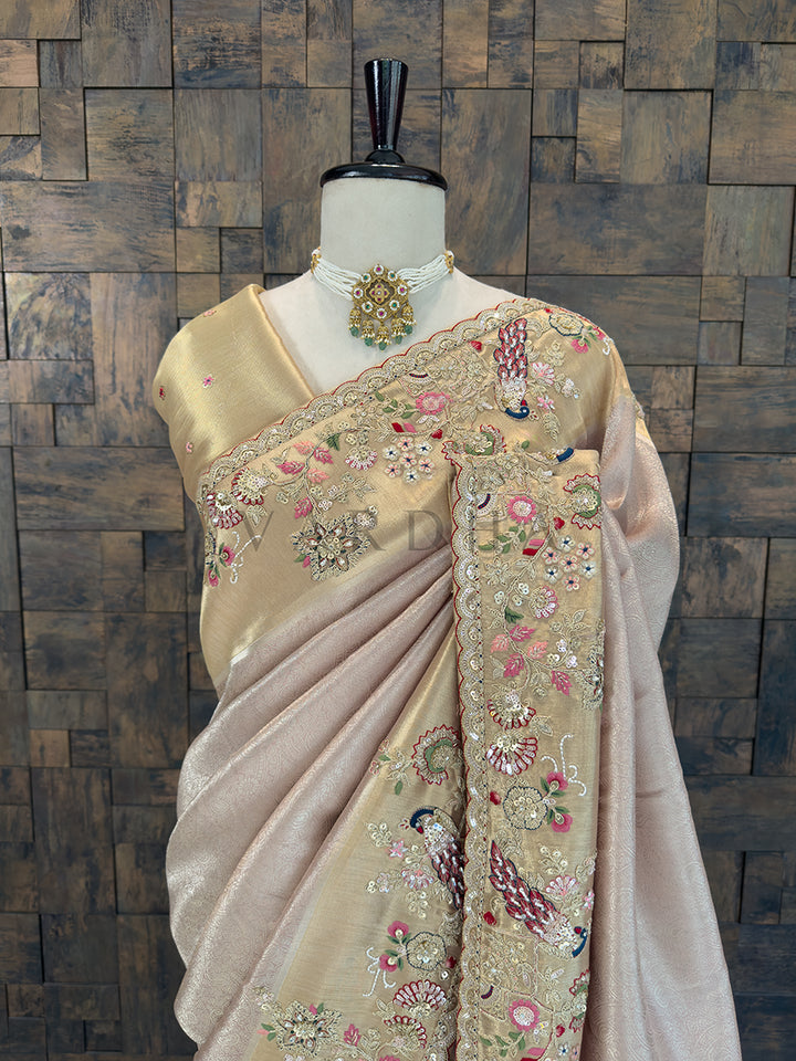Beige saree with floral embroidery on a mannequin against a wooden paneled wall. Close-up of a luxurious rose gold party-wear saree showing detailed zari, sequin, and thread work in floral and peacock motifs on the shoulder pallu, accessorized with a traditional pearl and kundan choker necklace.