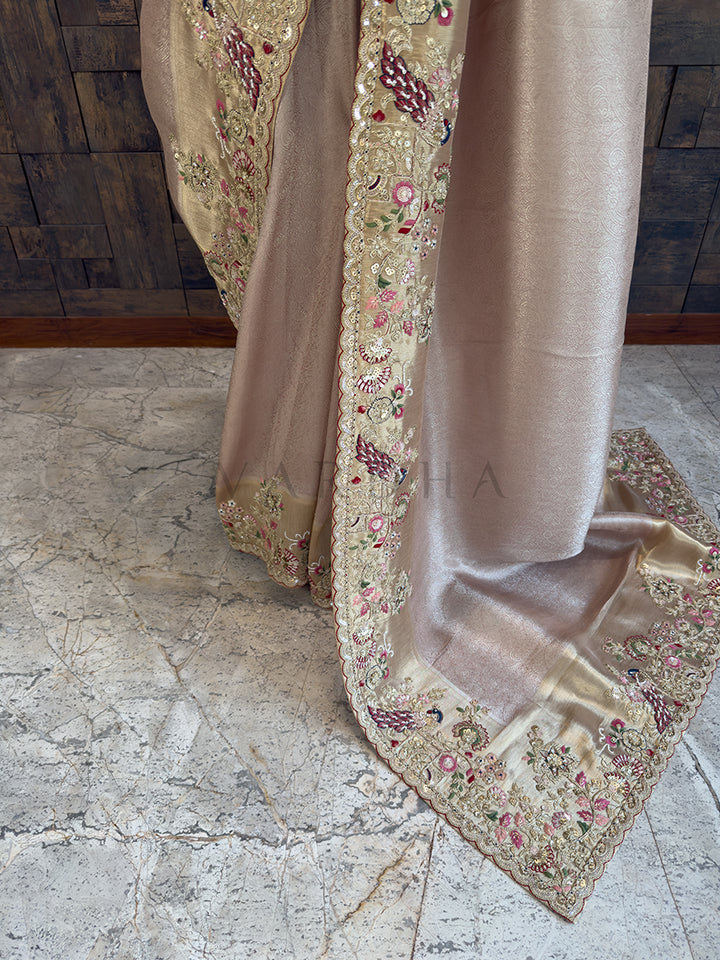 Beige saree with floral embroidery on a stone floor Lower drape of a shimmering beige silk ethnic saree highlighting the elegant fall and exquisite golden border, featuring colorful hand-embroidered peacock and floral designs cascading toward the floor.
