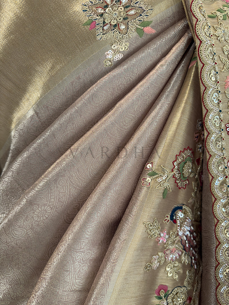 Close-up of a saree with intricate embroidery and patterns. Macro texture shot of a premium designer silk saree revealing subtle woven paisley patterns in the fabric pleats, contrasted alongside a heavily embellished gold border with rich pink and green floral embroidery.