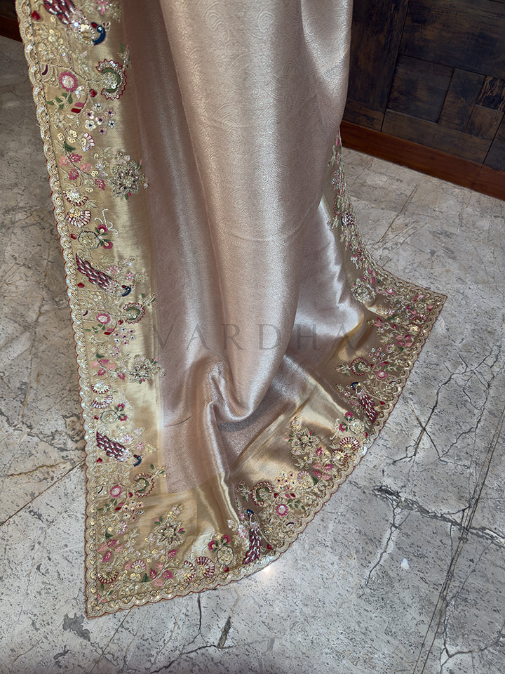 Silky fabric with floral embroidery on a textured floor Trailing edge of a metallic champagne bridal and guest-wear saree, displaying a heavy zardosi and sequin-embellished border with traditional Indian bird and floral motifs on a polished stone floor.