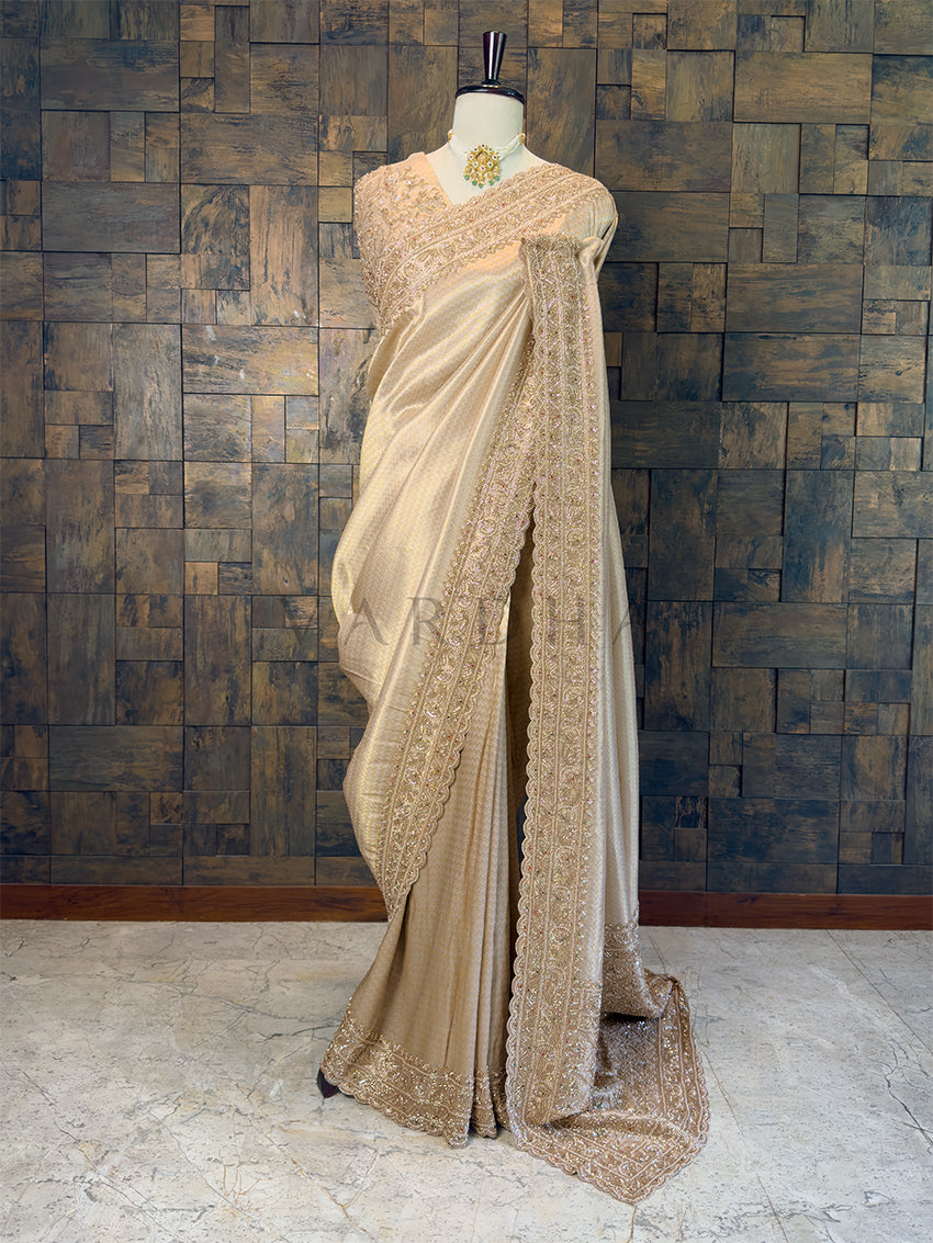 Cream saree with intricate patterns on a mannequin against a textured wall. Full View Chikoo Viscose Saree with Heavy Cutdana Handwork available with pre-drape services and free gift services.