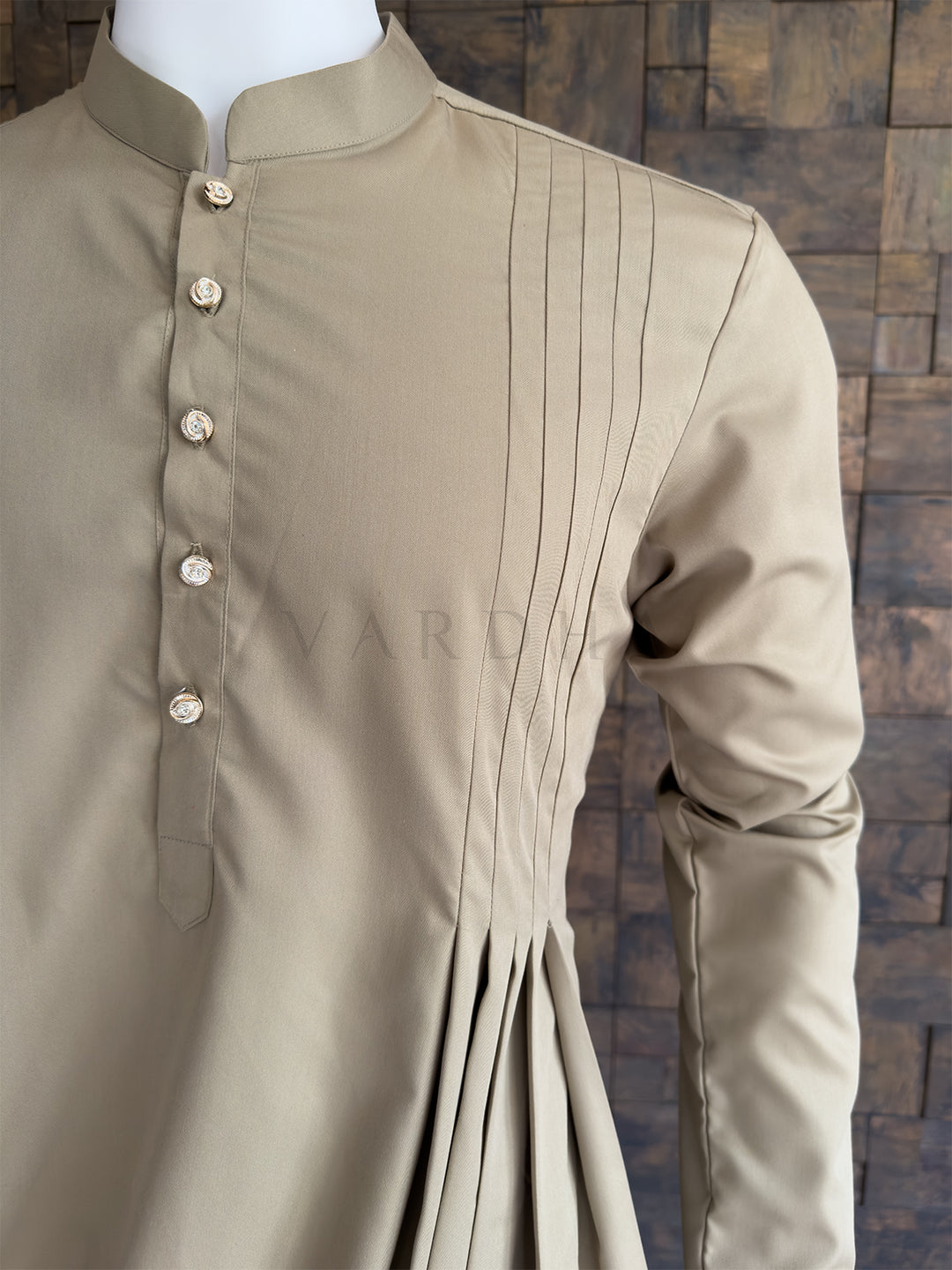Beige kurta with pleated details on a mannequin against a wooden paneled wall. Side angle highlighting the sharp pintucks flowing into the luxurious asymmetrical cowl drape on the premium cotton fabric of this stylish men&