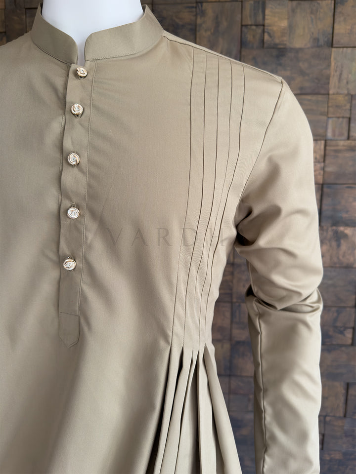 Beige kurta with pleated details on a mannequin against a wooden paneled wall. Side angle highlighting the sharp pintucks flowing into the luxurious asymmetrical cowl drape on the premium cotton fabric of this stylish men&