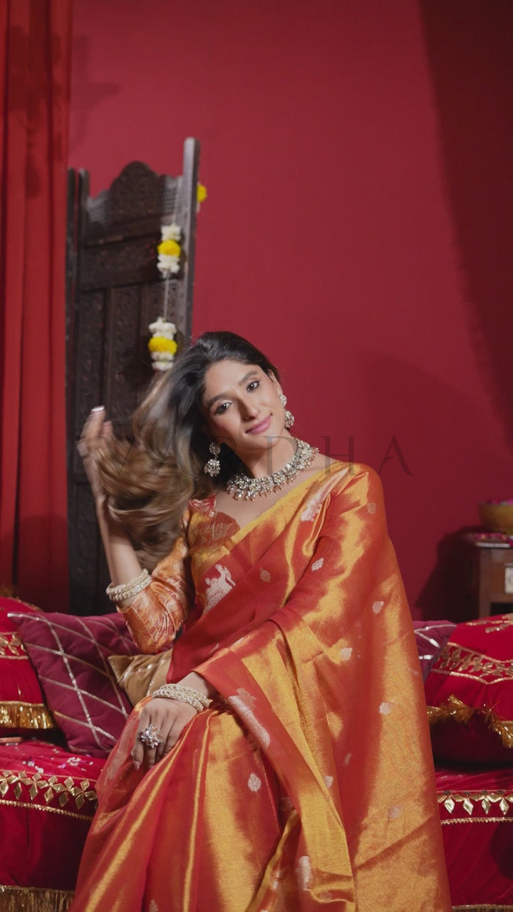 A woman wearing a rich orange and gold silk saree with traditional Indian jewelry, seated on a red upholstered sofa against a deep red wall. Glamorous shot of a woman flipping her hair, wearing a festive sunset orange heavy tissue silk saree perfect for weddings.
