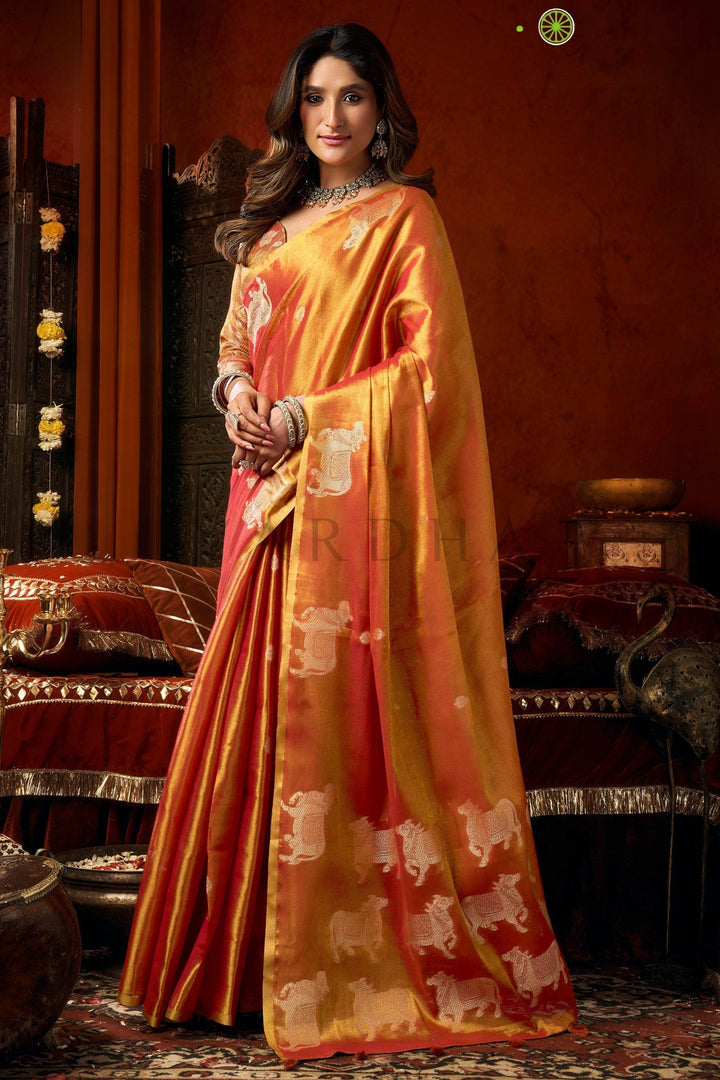 Woman wearing a traditional orange saree with white patterns in an elegant setting. Side standing view of the dual-tone red festive wear heavy tissue saree highlighting the traditional Pichwai-style motifs and flawless drape.