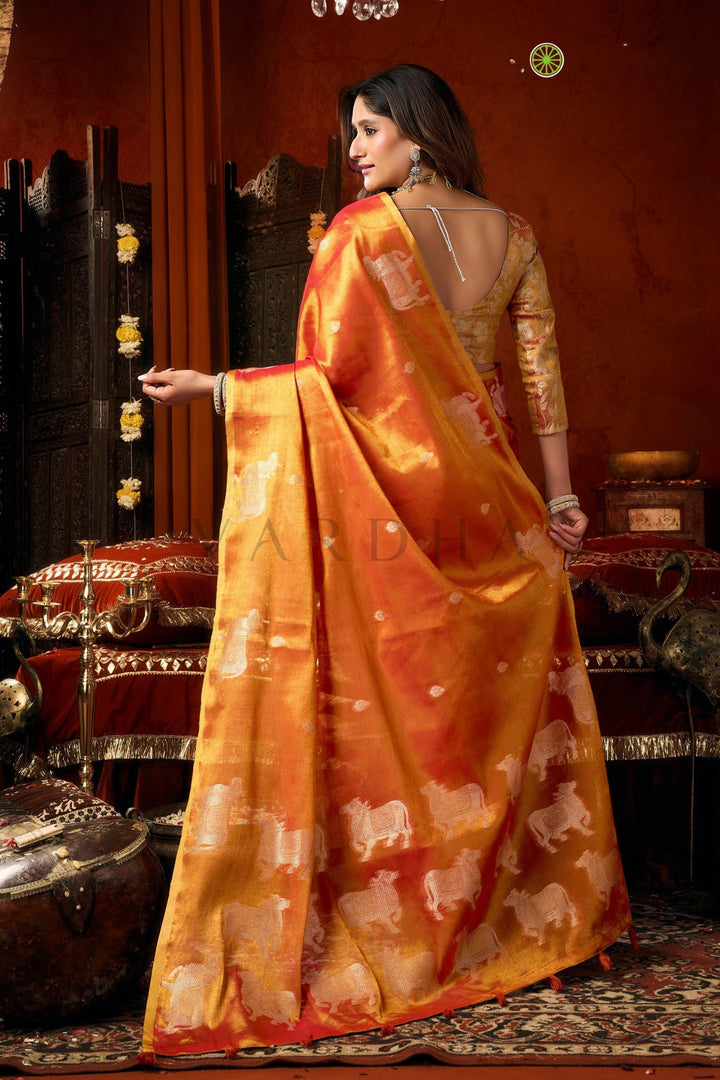 Woman in an orange saree with elephant patterns in a decorated room. Back view of the radiant coral red tissue saree, emphasizing the rich golden metallic sheen, woven silver cow designs, and elegant fancy tassels.