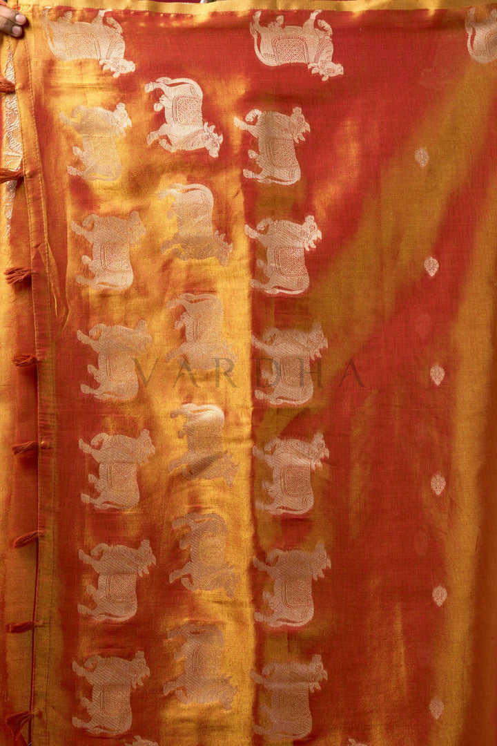 A close-up of a tie-dye fabric, likely a saree, featuring vertical stripes of orange, yellow, and red with white elephant motifs woven or printed throughout. Close-up of a beautiful woman wearing a sunset orange tissue silk saree with a matching woven floral blouse, perfect for festive occasions.