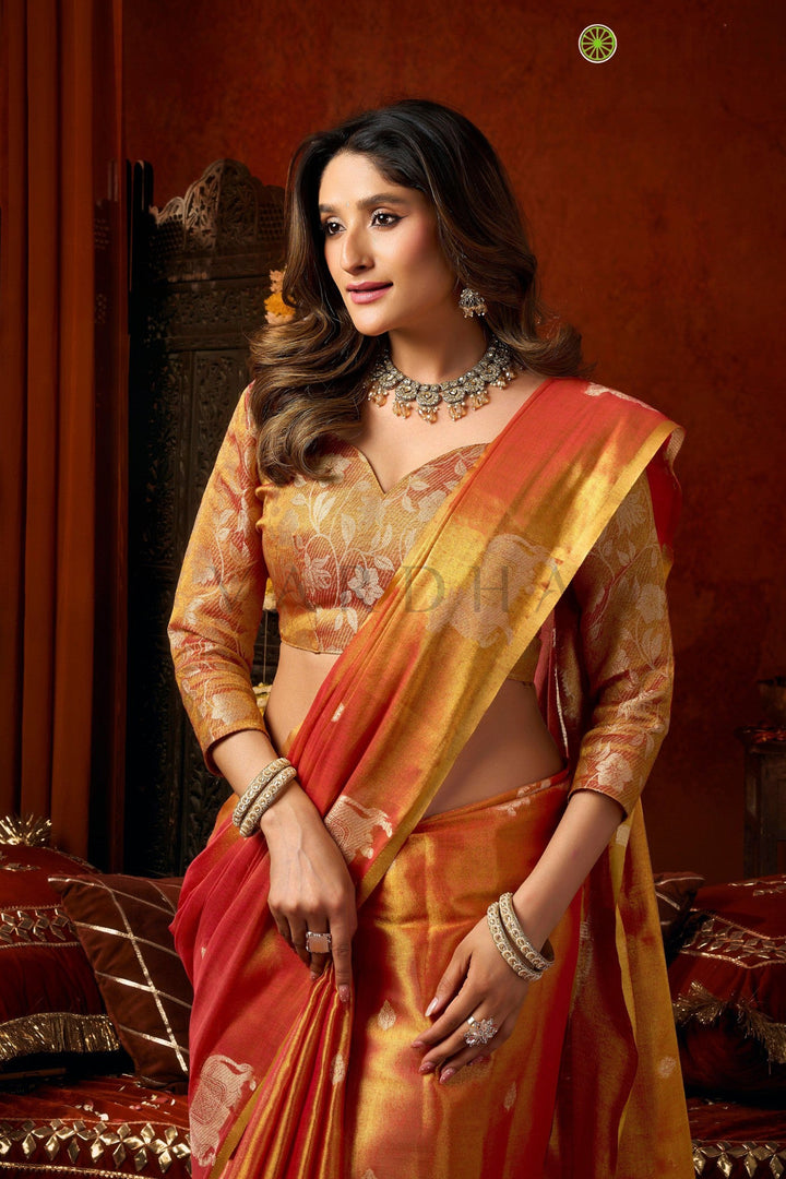 Woman in traditional orange saree with jewelry against a warm-toned background Elegant pose of a woman adjusting her earrings while wearing the dual-tone sunset orange tissue silk saree for a festive celebration.