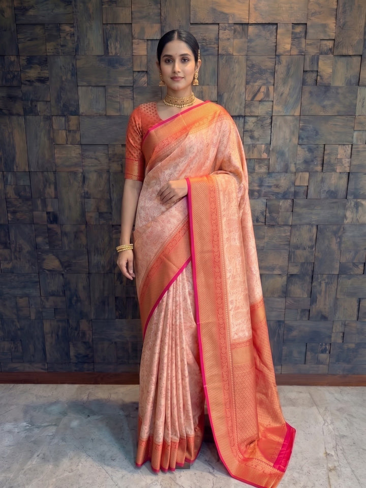 A woman wearing a peach and orange saree with a bright pink border, standing in front of a dark textured stone wall. A full-length front view of a woman elegantly modeling a cream-red Tanchoi satin silk saree. She has her hands softly resting near her waist, highlighting the intricate zari work on the rich red border that beautifully frames the subtle cream body of the draped fabric.