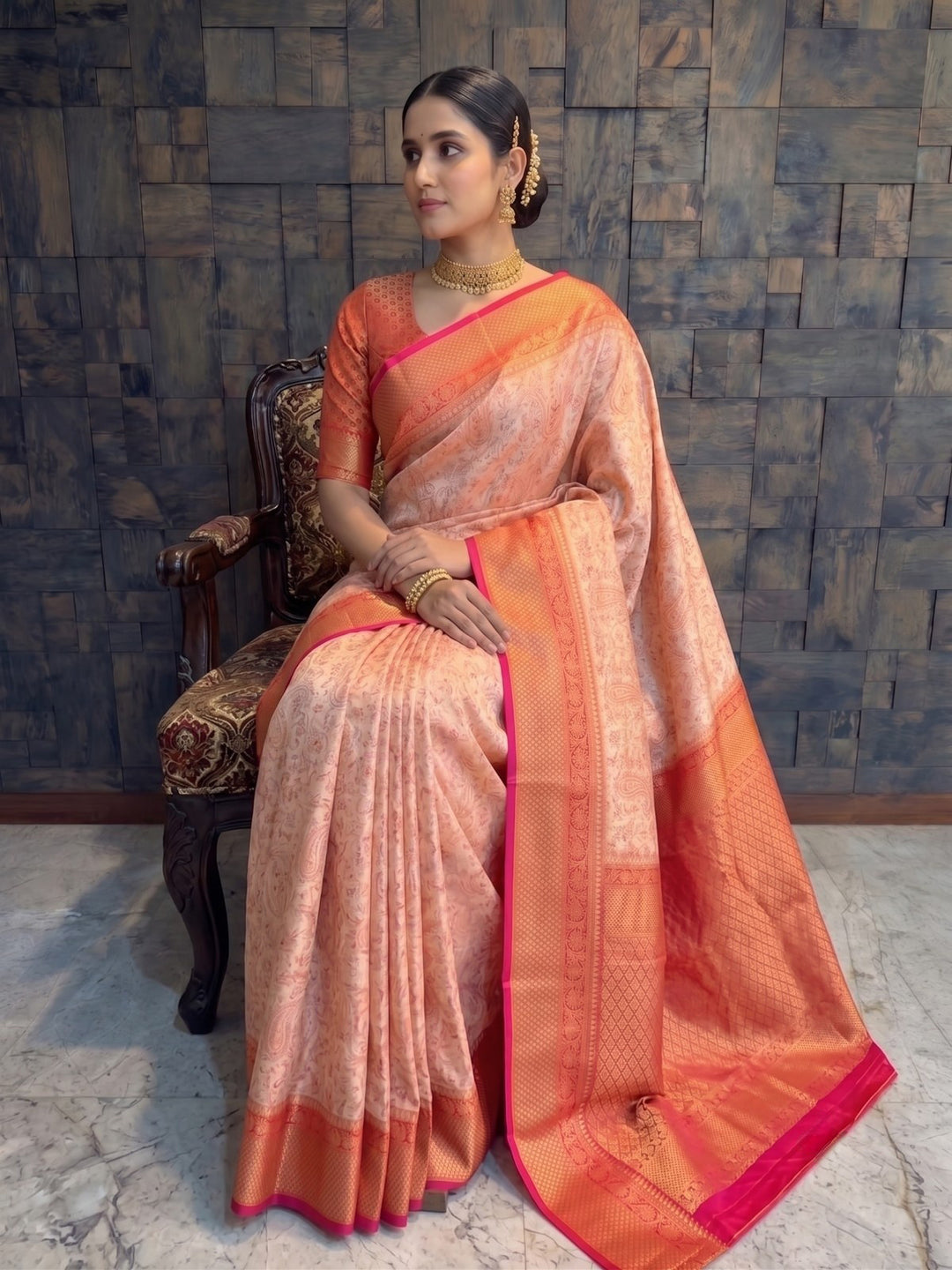 A woman seated on a dark wooden chair wearing a peach and orange silk saree with a bright pink border, posed against a textured gray stone wall. A full-length portrait of a woman seated gracefully on an ornately carved wooden armchair with patterned upholstery. She is dressed in the cream-red Tanchoi satin silk saree, with her hands resting gently on her lap and the rich red zari pallu draped beautifully over the side of the chair toward the floor.