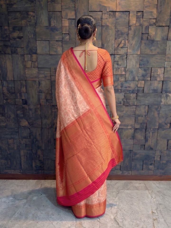 A woman viewed from behind wearing a peach and orange saree with a bright pink border and a matching orange blouse, standing against a dark textured stone wall. A full-length back view of a woman draped in a cream-red Tanchoi satin silk saree. The shot highlights the elegant back design of her matching reddish-orange blouse, which features delicate string ties, alongside the heavy gold zari weaving on the vibrant red pallu cascading down her left shoulder.