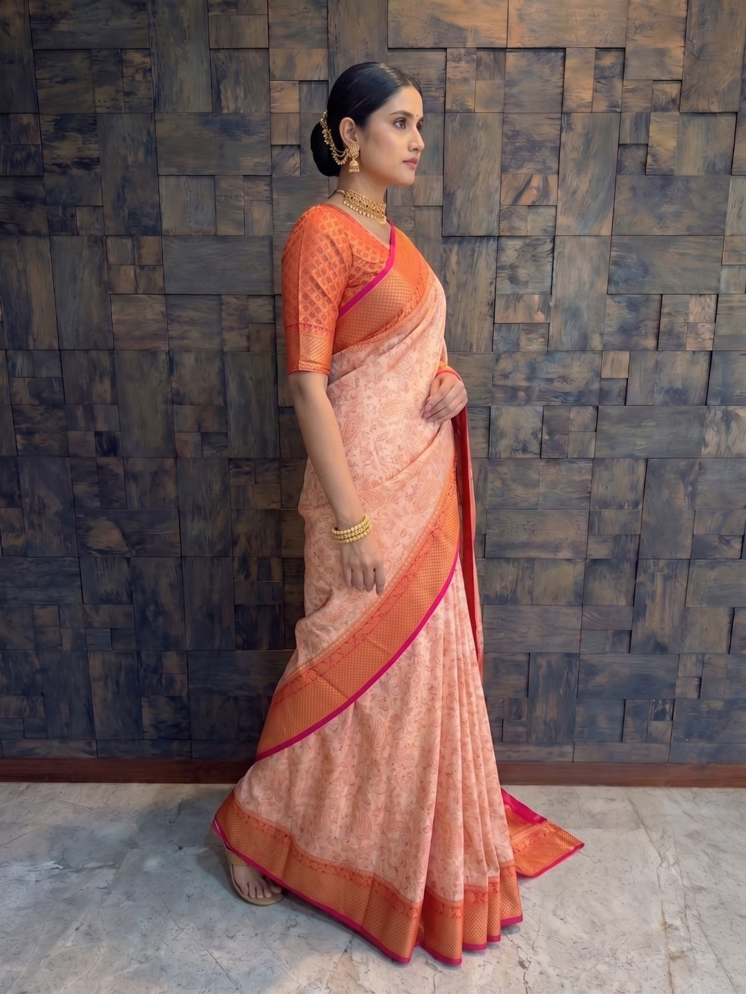 A full-length side profile view of a woman stepping slightly forward in a cream Tanchoi satin silk saree. The side angle beautifully showcases the continuous contrasting red and gold zari border outlining the elegant drape of the cream fabric against the dark wooden background.
