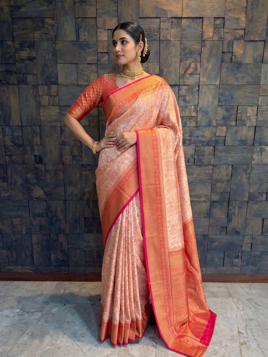 A full-length front view of a woman confidently posing with her right hand resting on her hip, wearing a cream Tanchoi satin silk saree. The dynamic pose emphasizes the crisp front pleats and the thick red and gold zari border wrapping around her waist and draped evenly over her shoulder.
