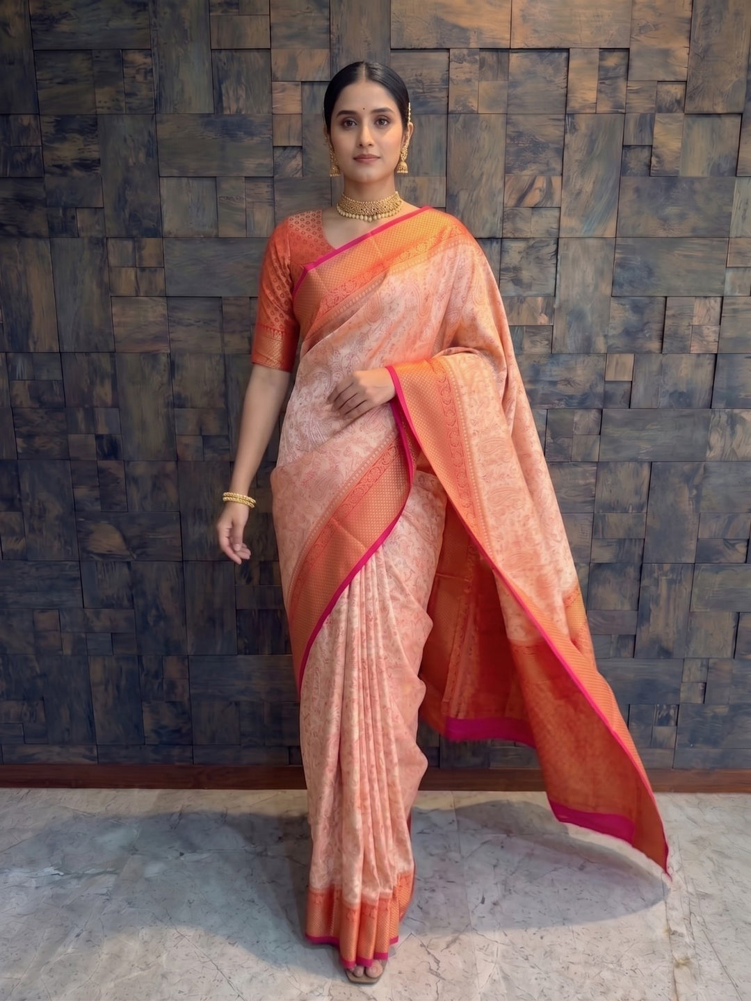 A woman modeling a peach and orange silk saree with a pink border, standing against a dark textured wooden wall. A full-length front view of a woman posing in a cream Tanchoi satin silk saree featuring subtle woven body motifs and a vibrant red and gold zari border. She is wearing a matching reddish-orange patterned short-sleeved blouse and gold jewelry, standing against a textured wooden paneled backdrop with the red pallu flowing gracefully to her side.