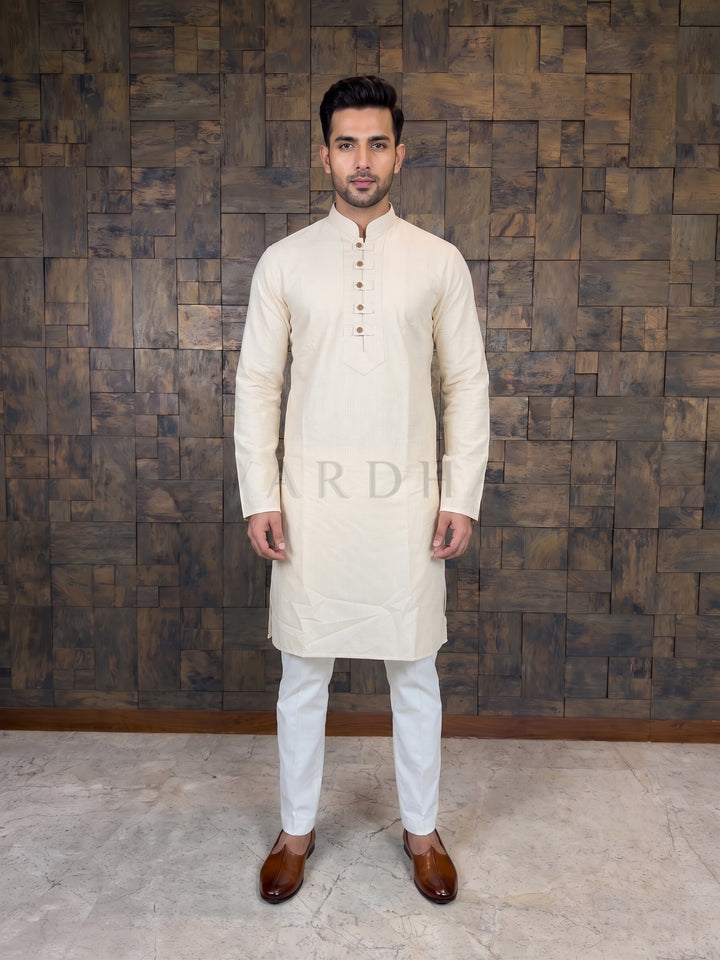 Man wearing a cream kurta with white pants against a wooden paneled wall. Cream Designer Cotton Kurta. Designed for the modern gentleman who values impeccable tailoring over heavy embellishments